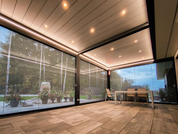 Überdachte Terrasse mit Glaswänden, beleuchtetem Deckenpaneel und einem Tisch mit Stühlen