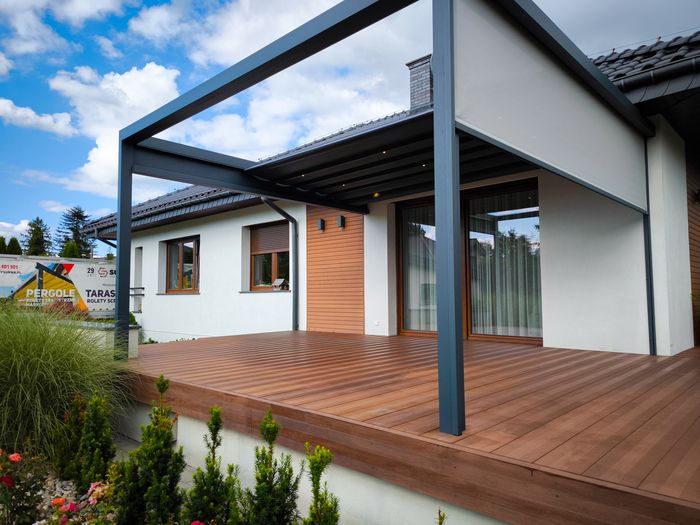 Moderne Terrasse mit Holzbelag und schwarzer Pergola vor weißem Haus mit Fenstern und Tür