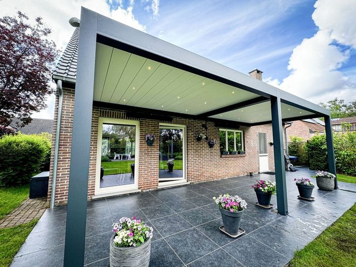 Backsteinhaus mit überdachtem Eingangsbereich auf dunklem Untergrund mit Topfpflanzen, grüner Umgebung und klarem Himmel