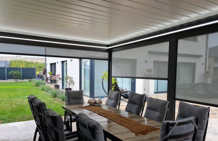 Eine überdachte Terrasse mit einem hölzernen Esstisch und Stühlen, umgeben von eingebauten grauen, leicht abdunkelnden Rollos vor einem Haus mit Rasen