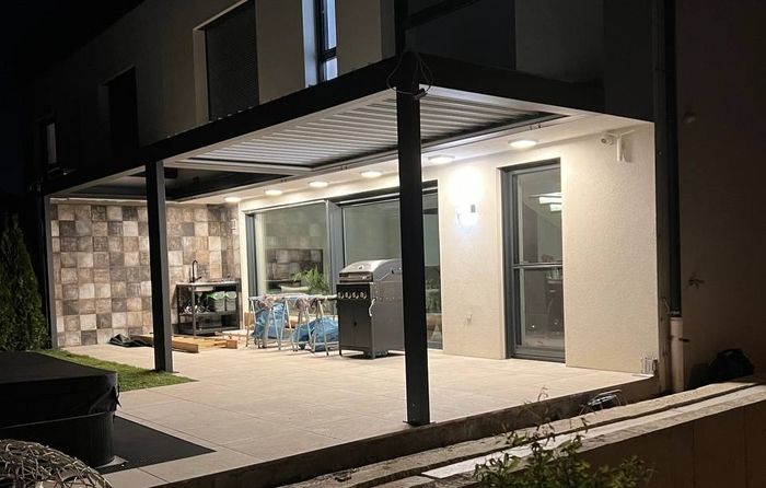 Nachtansicht einer modernen Terrasse mit Grill, einem mit Plastik abgedeckten Essbereich und einer Steinmauer. Der Bereich wird von Deckenleuchten beleuchtet, wodurch eine gemütliche und einladende Atmosphäre entsteht.