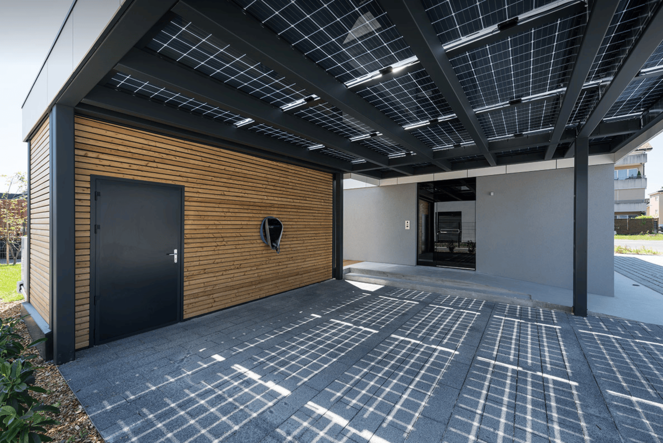 Carport mit Dach aus Solarpaneelen, daneben Holzverkleidung mit schwarzer Tür und Ladestation für Elektroautos