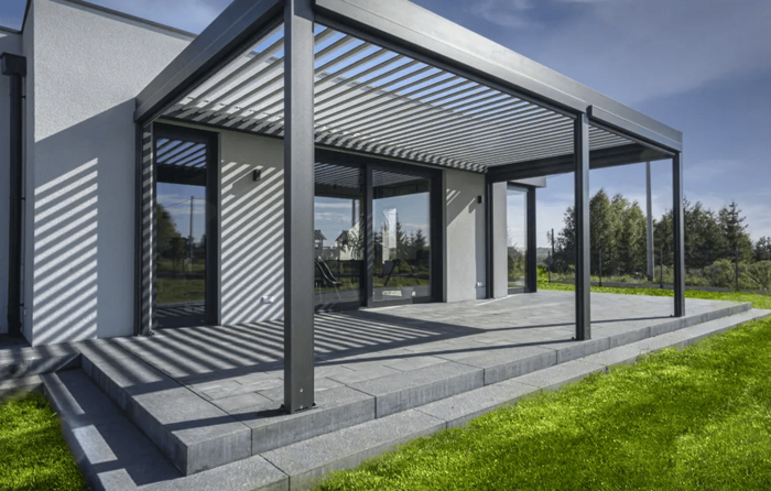 Moderne Terrasse mit grauer Pergola aus Metall und Glasfront eines Hauses, umgeben von grünem Rasen