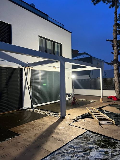 Moderner weißer zweistöckiger Wohnhaus mit großen Fenstern und einer überdachten Terrasse bei Dämmerung, Baustelle mit Leiter und Abdeckungen im Vordergrund