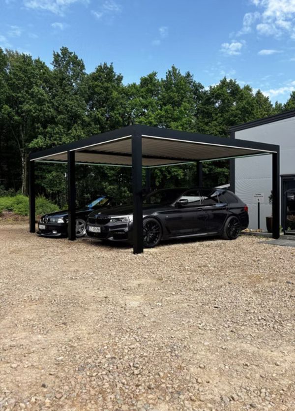Zwei elegante schwarze Autos stehen unter einem modernen, metallischen Carport neben einem weißen Gebäude. Im Hintergrund sind üppig grüne Bäume und ein blauer Himmel zu sehen.
