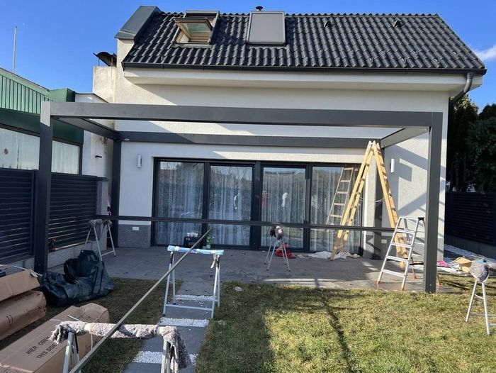 Ein Haus, an dem gerade eine Terrasse gebaut wird, mit einem teilweise montierten schwarzen Metallrahmen. Eine Leiter und Werkzeuge liegen verstreut herum. Sonnig, mit klarem blauem Himmel.