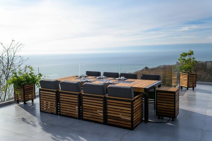 Terrassengarnitur mit Holztisch und gepolsterten Stühlen auf einer gefliesten Terrasse. Umgeben von Glasgeländer, Pflanzen und Meerblick unter blauem Himmel.