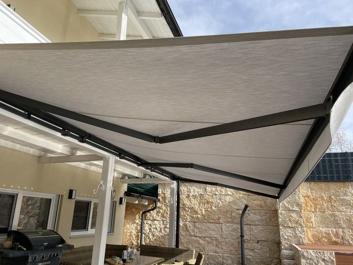 Eine große beigefarbene Markise spendet Schatten auf einer Terrasse mit Korbmöbeln und einem Grill. Eine Steinmauer und Fenster schaffen einen gemütlichen Außenbereich.