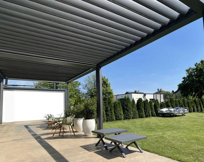 Überdachte Terrasse mit moderner Lamellendachkonstruktion, Gartenstühlen und großen weißen Pflanzgefäßen, Blick auf gepflegten Rasen und Hecke