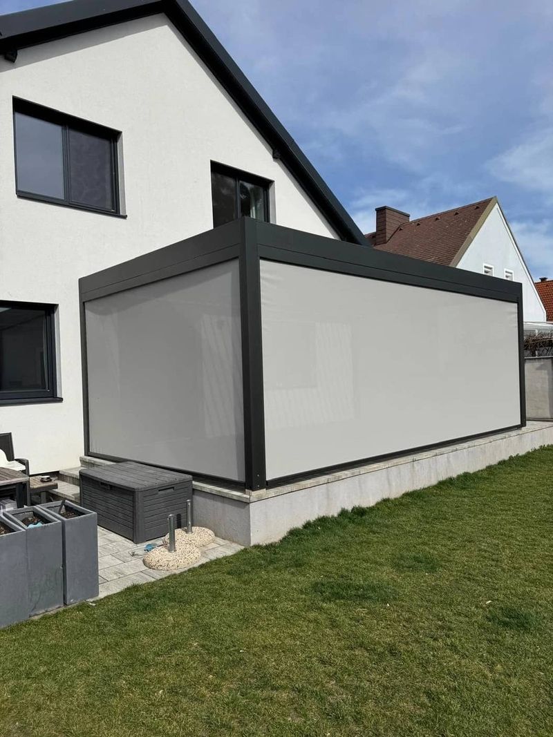 Eine moderne graue Terrassenüberdachung, die an ein weißes Haus angebaut ist, mit großen Fenstern und einem gepflegten Rasen.