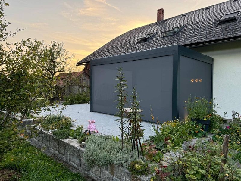 Eine moderne graue Terrassenüberdachung neben einem Haus, umgeben von bunten Gartenpflanzen und Blumen, unter einem Abendhimmel.