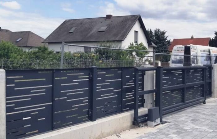 Moderner schwarzer Metallzaun mit horizontalen Schlitzen vor einem weißen Einfamilienhaus mit grauem Dach