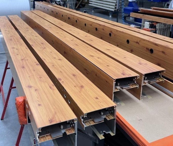 Holzähnliche Metallträger, die auf einem Werkstatttisch ausgerichtet sind und eine hellbraune Oberfläche mit Maserung aufweisen. Industrielle Umgebung mit Werkzeugen im Hintergrund.