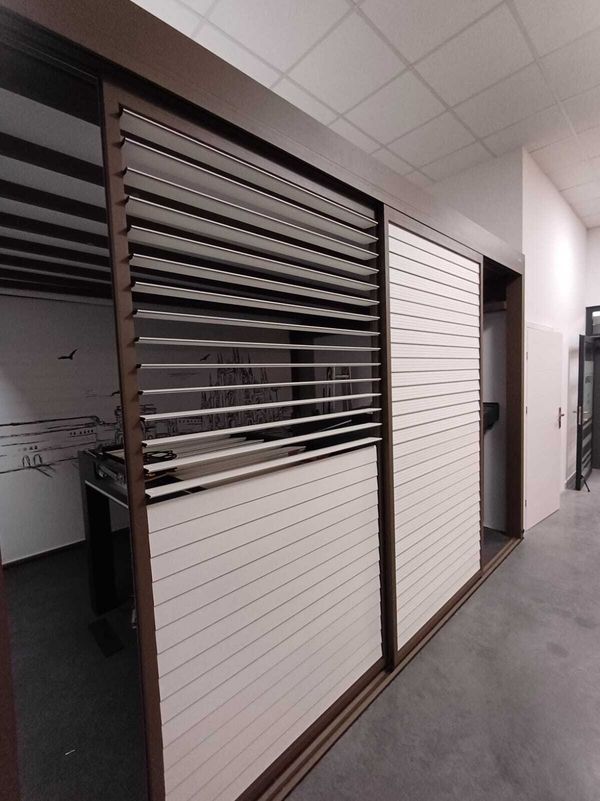 Innenraum eines modernen Büros mit braunen und weißen Wandpaneelen. Die Jalousien sind teilweise geöffnet und geben den Blick auf einen Arbeitsbereich mit Wandkunst frei. Die Umgebung ist ruhig und strukturiert.