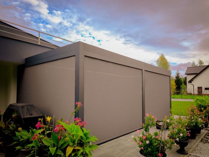 Moderne Terrasse mit eleganter grauer Pergola und farbenfrohen Topfpflanzen auf Fliesenboden. Der bewölkte Himmel sorgt für eine ruhige, stilvolle Atmosphäre.