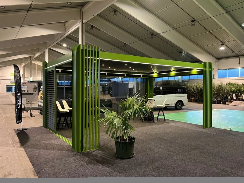 Moderner grüner Messestand mit klaren Linien, beleuchteter Decke, Stühlen und einer Topfpflanze in einem großzügigen Veranstaltungsraum.