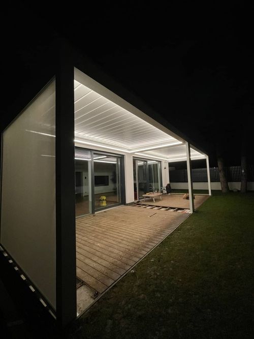 Moderner Innenhof bei Nacht, mit warmer Beleuchtung unter einer eleganten, weißen Pergola. Die Holzterrasse führt zu Glastüren, an deren Seite sich eine Rasenfläche befindet.