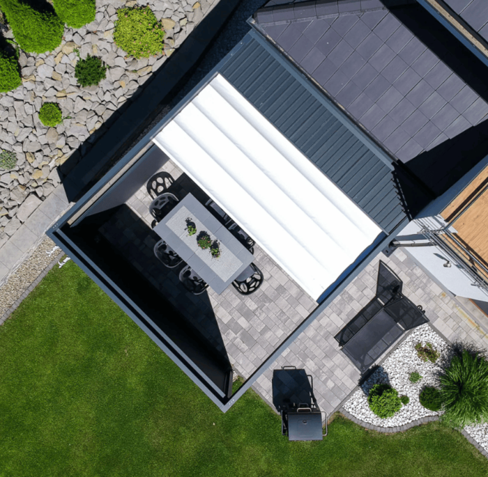 Luftaufnahme einer modernen Terrasse mit einer weißen Pergola, einem grauen Tisch und Stühlen. Der umgebende grüne Rasen und die dekorativen Steine sorgen für eine frische, ruhige Atmosphäre.
