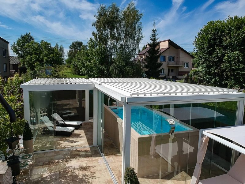 Eine moderne, verglaste Terrasse mit Swimmingpool und Liegestühlen, umgeben von üppigem Grün und blauem Himmel.