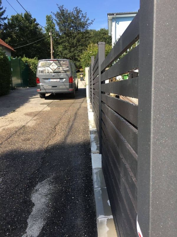 Grauer Metallzaun entlang eines Gehwegs mit einem geparkten grauen Van im Hintergrund unter Bäumen