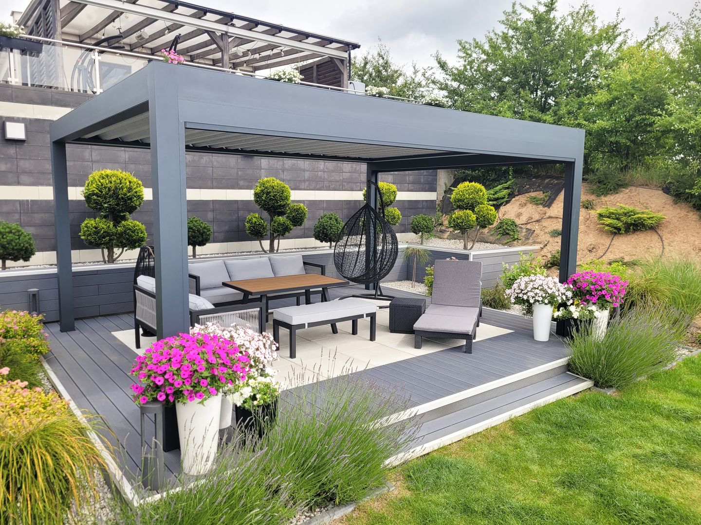 Moderner Gartenpavillon mit grauer Metallstruktur, Sitzgruppe mit Hängesessel, Liegestuhl und Tisch auf Holzterrasse, umgeben von grünen Pflanzen und bunten Blumen