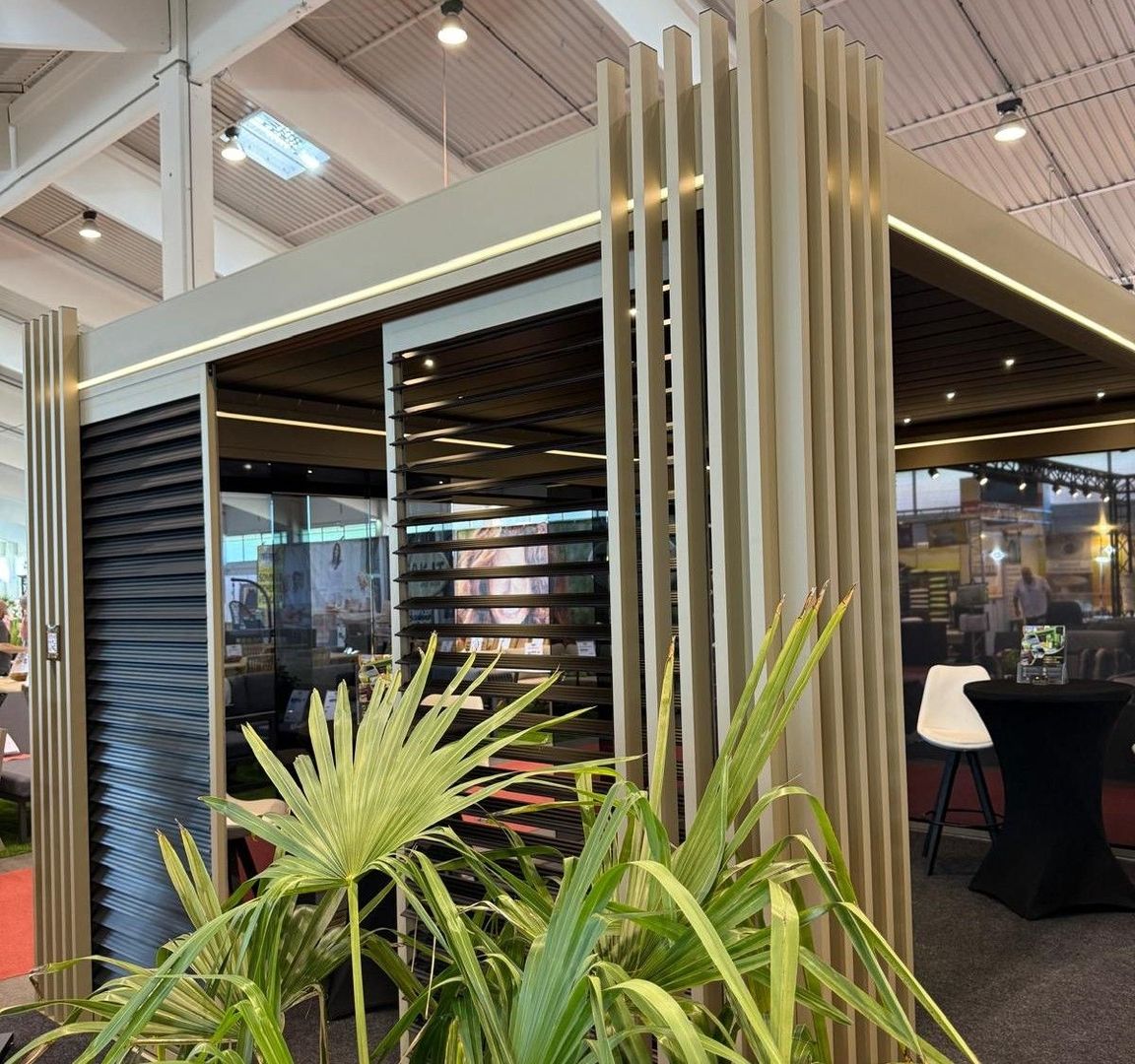 Beigefarbener, überdachter Messestand mit einem Tisch, Stühlen und mehreren Pflanzen dekoriert.